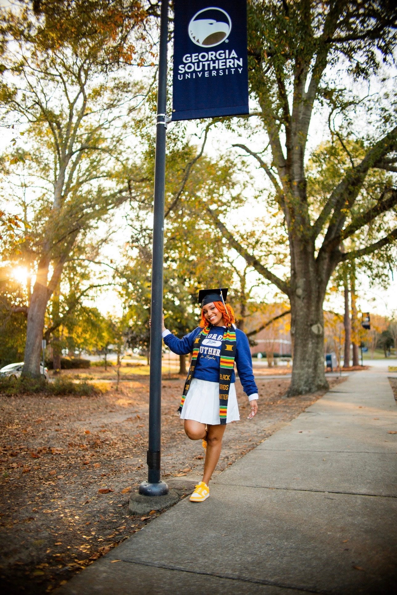 Taylor Pledger in a white skirt, blue Georgia Southern sweatshirt, yellow sneakers, graduation cap and stole. She is holding a pole with a blue Georgia Southern banner. Photo Credit: Gregory Martin