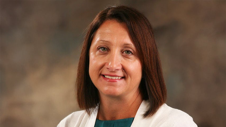 Portrait of a smiling woman with shoulder-length brown hair wearing a white blazer over a teal blouse, against a neutral studio backdrop.