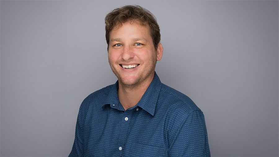 Smiling man with short brown hair in a blue button-down shirt against a gray backdrop.