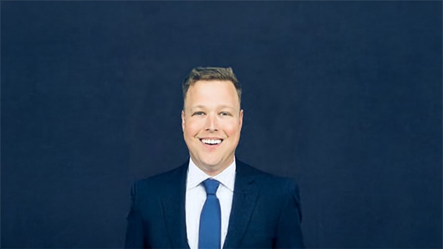 Professional headshot: smiling man in a navy suit and blue tie against a dark blue backdrop, suitable for business profiles.