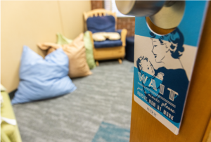 A room with comfortable seating and pillows on the floor, A sign on the door warns visitors of lactation in progress.