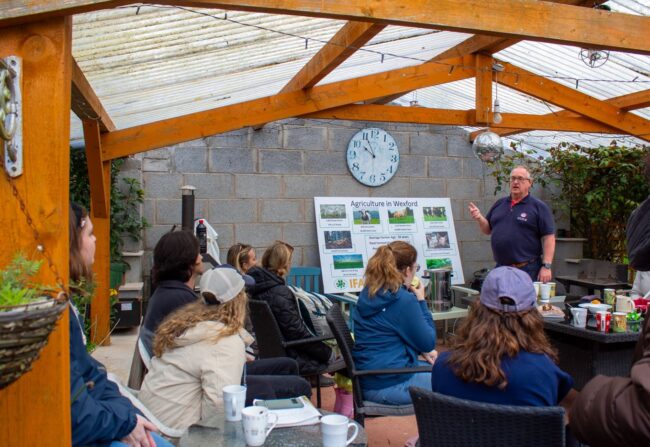 Instructor explains a poster titled 'Agriculture in Wixford' to a seated audience inside a wooden-framed glasshouse.