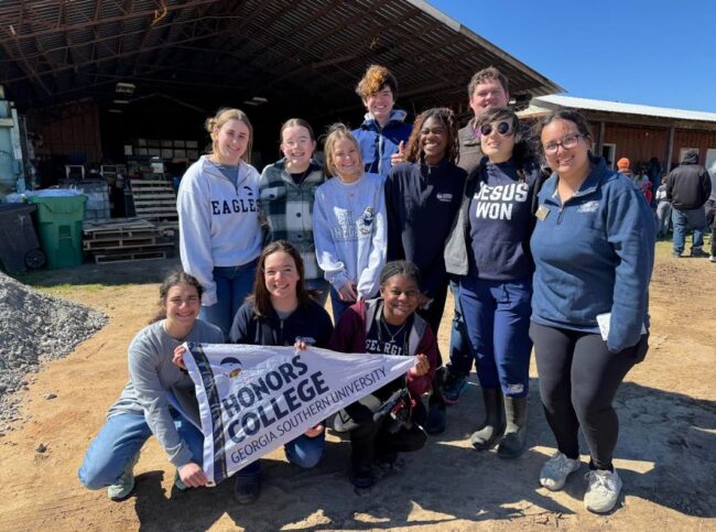 Group of smiling students posing outdoors, holding a banner that reads 'Honors College, Georgia Southern University' in front of a barn-like building.