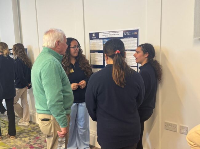Group of students explain a research poster to an older man in a hallway at a conference or event.