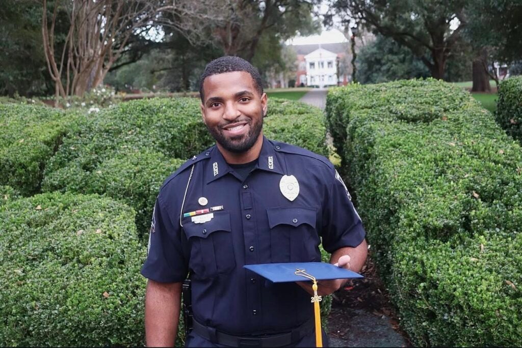 University Police Investigator Heiko Ellis stands in front of the GSU bushes on Sweetheart Circle at Georgia Southern University. He is wearing his police uniform and holding his 2025 grad cap and tassel.
