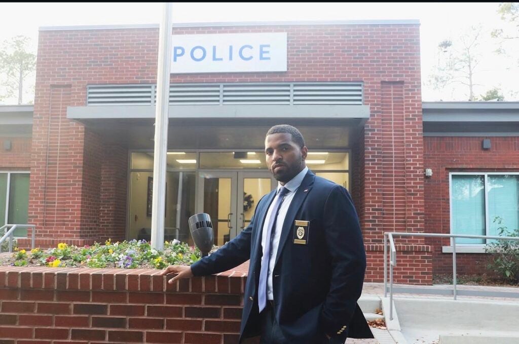 University Police Investigator Heiko Ellis stands in front of the University Police Department at Georgia Southern University. He is wearing a dark blue suit and his badge.