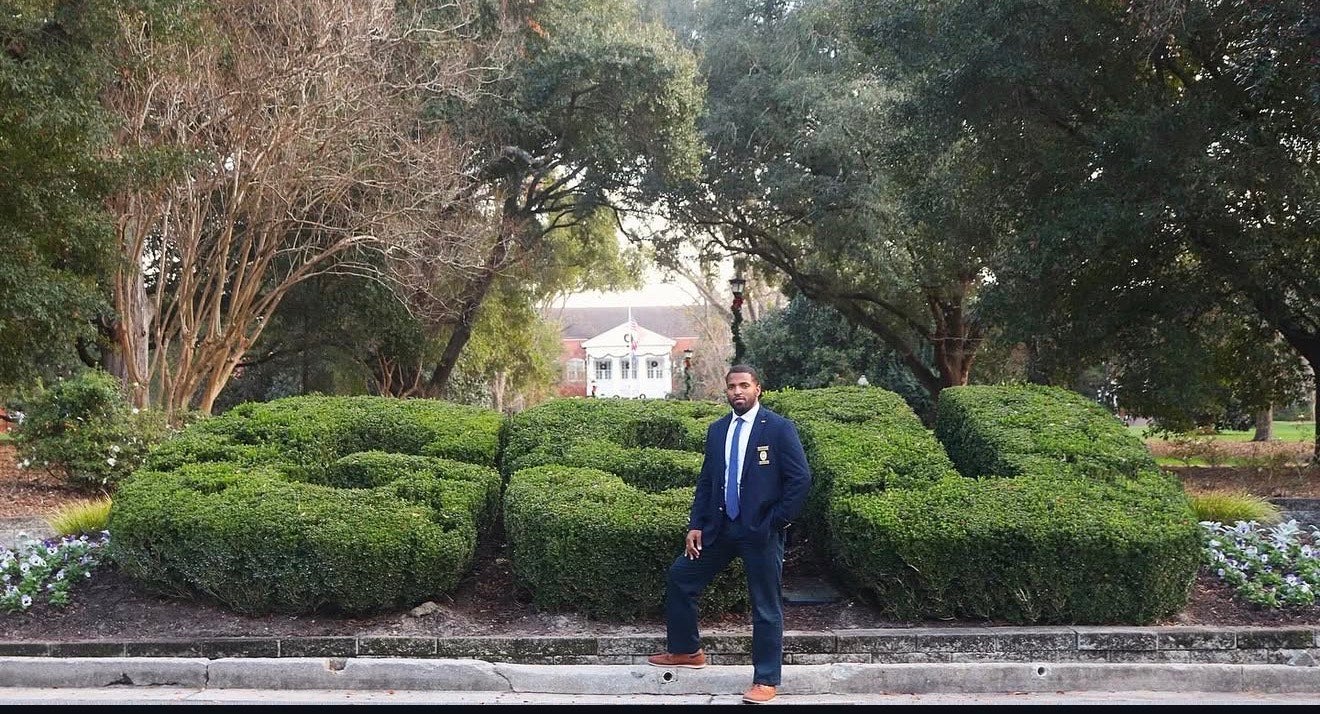 University Police Investigator Heiko Ellis stands in front of the GSU bushes on Sweetheart Circle at Georgia Southern University. He is wearing a dark blue suit and his badge.
