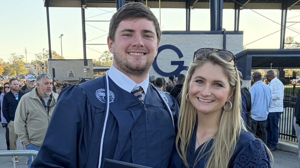 Man and woman pose together at graduation with man in graduation wear and his wife to his left side in a blue ruffled dress