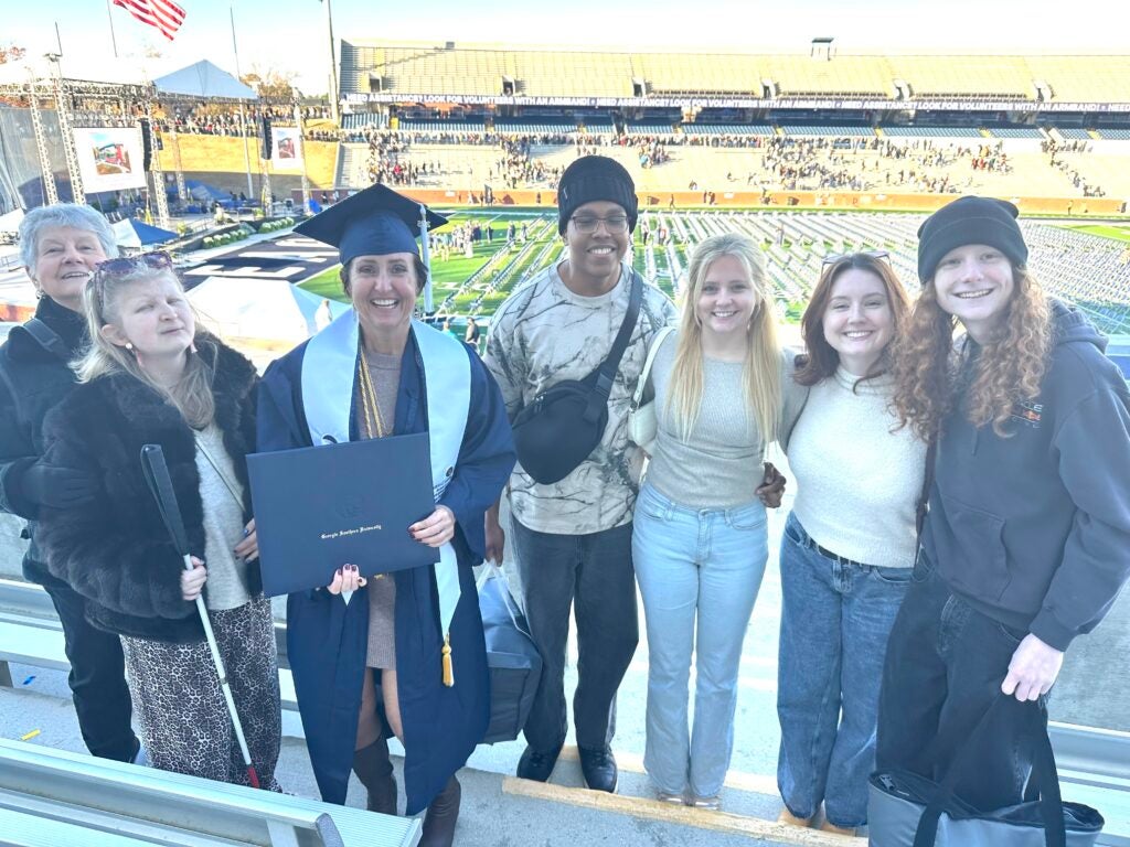 Georgia Southern December 2025 graduate poses with six supportive loved ones in the stands at Paulson Stadium. The woman graduating holds her diploma.