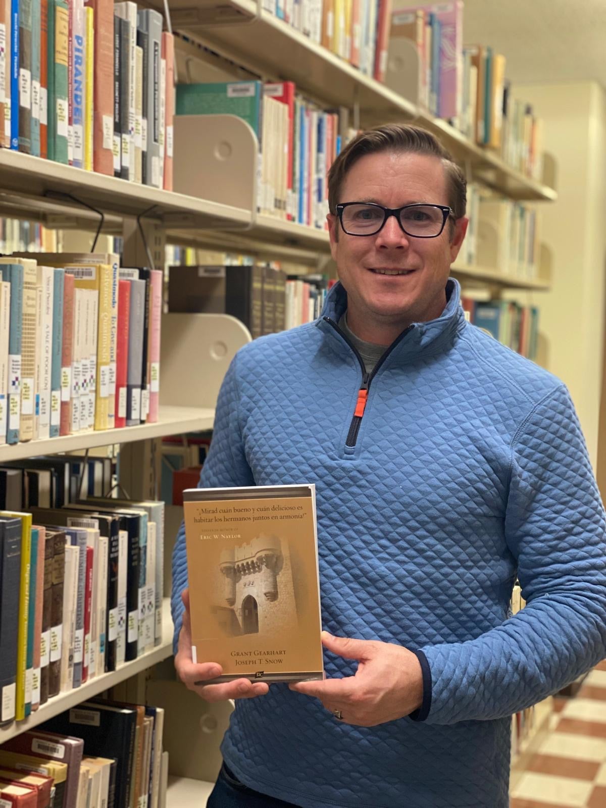 Professor Grant Gearhart holds a book while standing next to a library shelf full of books