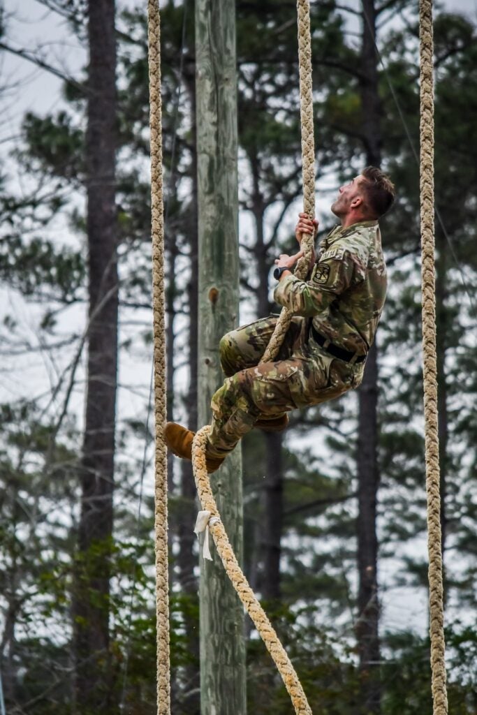 Georgia Southern Eagle Battalion ROTC male cadet climbs a rope in camoflage fatigues looking up at the sky with a forest behind him