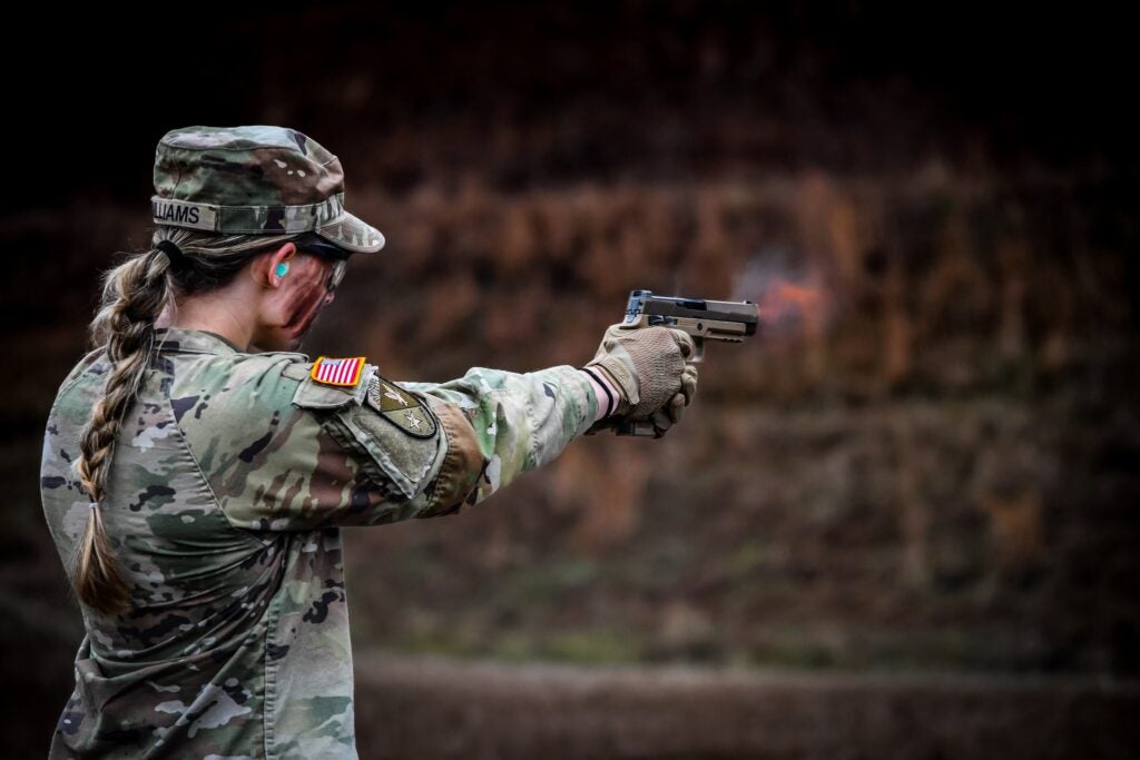 Female soldier with ponytail holds gun pointing at target in camoflage fatigues wearing a camo cap with her ash-blonde hair in a low braided ponytail 