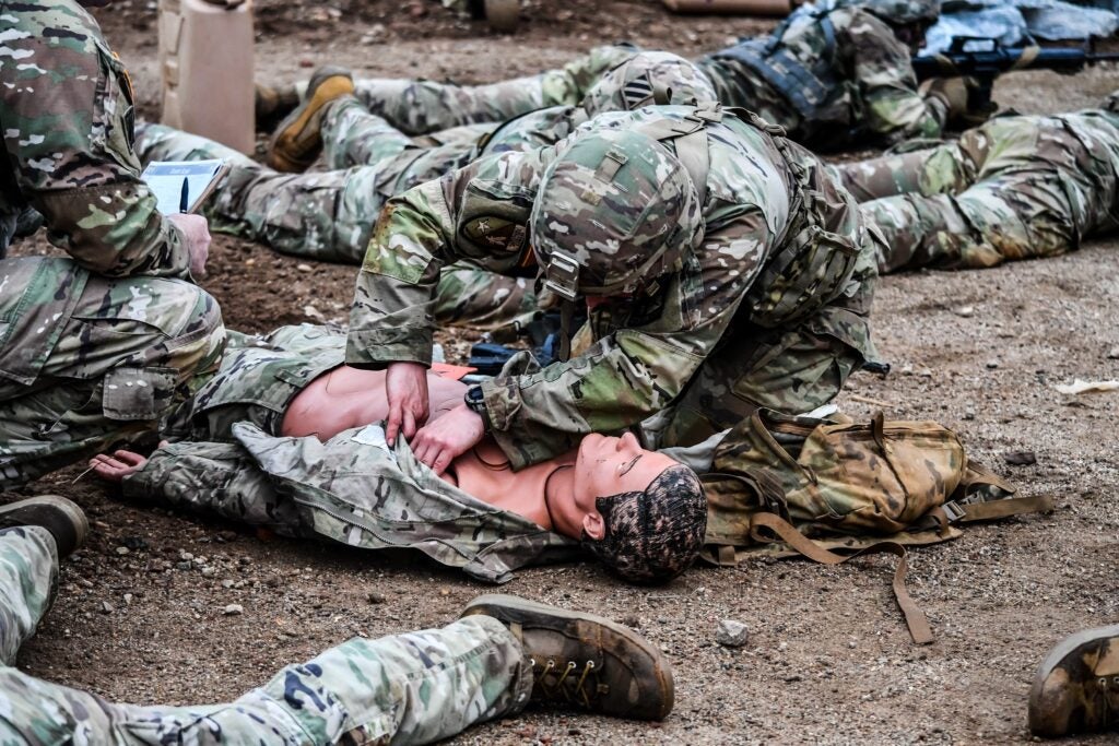 Eagle Battalion cadet in camouflage fatigues performs a medical procedure on a mannequin also dressed in camo fatigues. The cadet is wearing a helmet and hovering over the dummy.