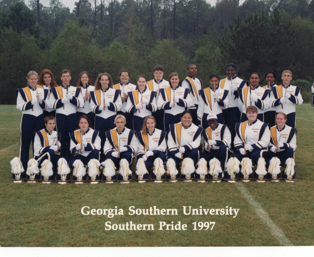Southern Pride flute section students pose together in white, gold and blue marching band uniforms with their white feather plumed hats lined up in front of them on the football field. 