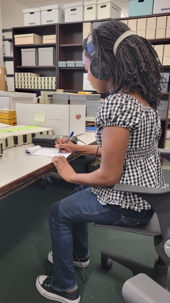 Ariana sits at a table and listens to headphones while transcribing content onto a notepad