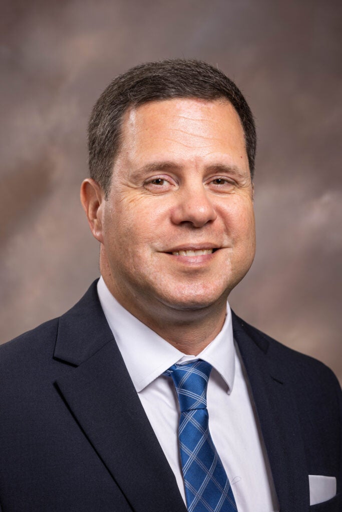 Joe Kardouni smiles while wearing a dark suit, white shirt and blue tie