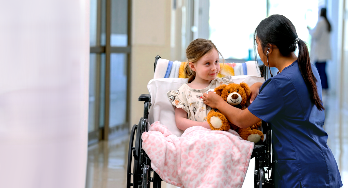 Georgia Southern nursing student in a hospital kneeling next to a young girl in a wheelchair, holding a teddy bear. The student is listening to the young girl's heart beat with a stethoscope.
