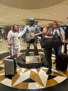Three students carrying luggage pose next to a statue of Elvis