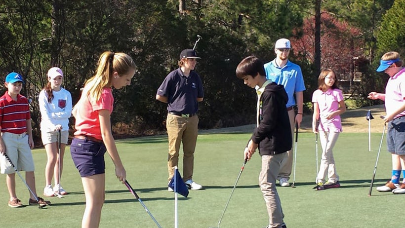 A group of students practices putting on a sunny golf green under the guidance of instructors.