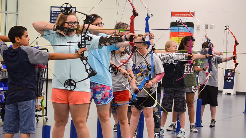 A group of students stands in a line indoors, focused on aiming their compound and recurve bows during an archery lesson.