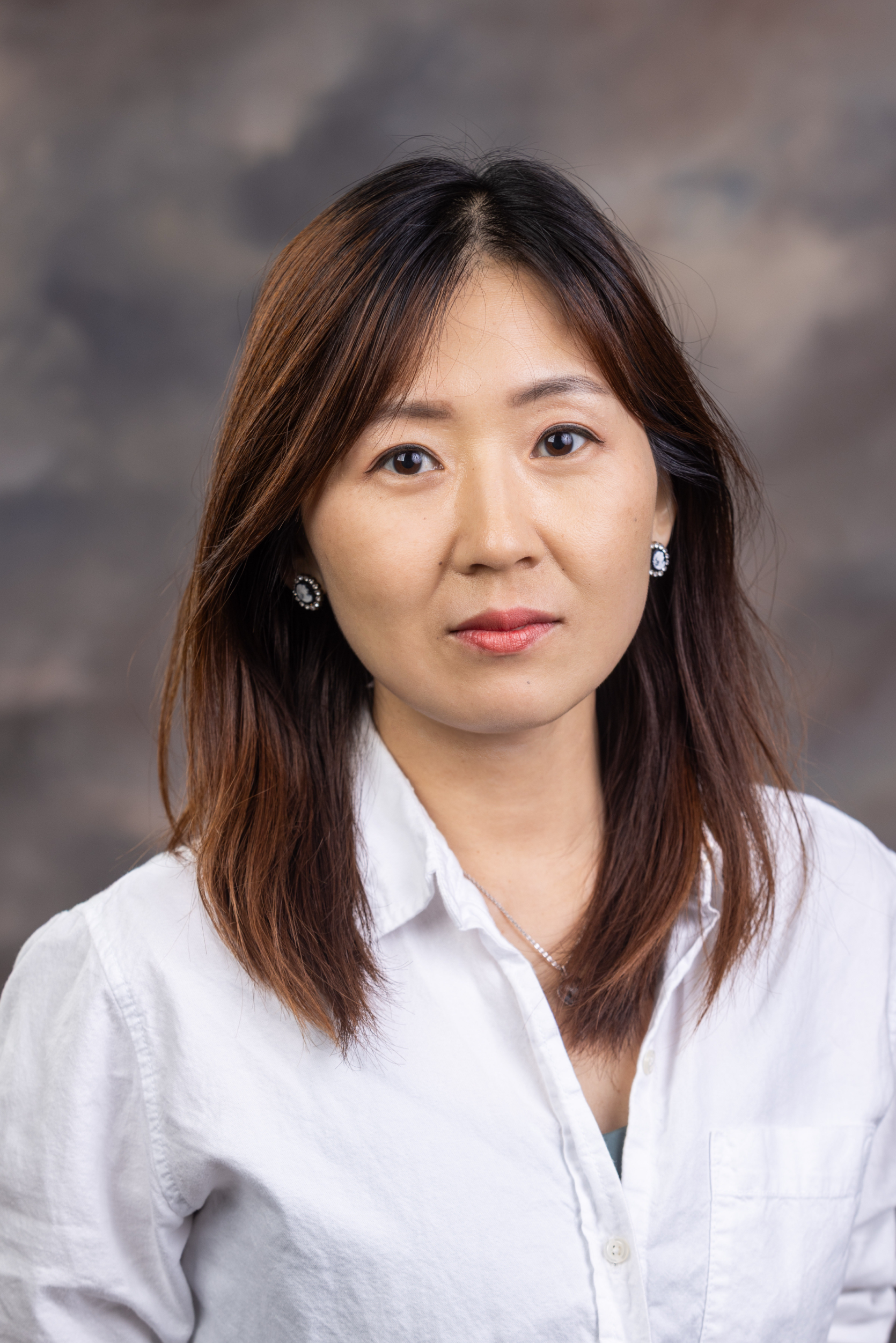 Close-up portrait of a woman with dark hair and brown highlights, wearing a white button-down shirt, against a neutral studio backdrop, looking at the camera.