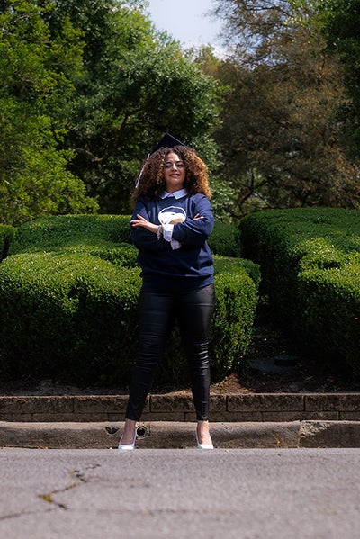 Graduate in a GSU tshirt standing with arms crossed on a path, in a park with trimmed hedges behind her.
