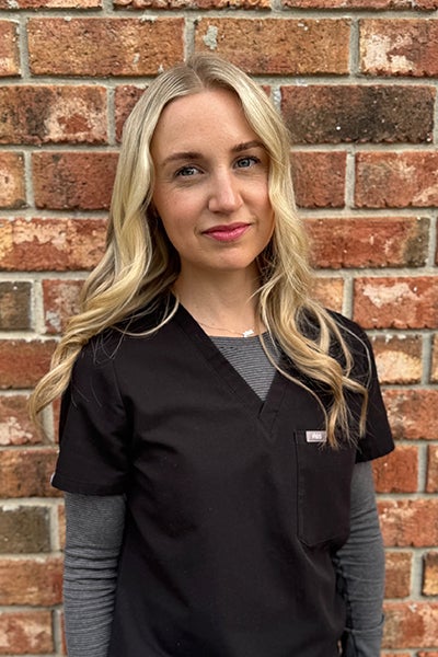 Woman with long blonde hair wearing black scrubs over a gray long-sleeve shirt, standing in front of a brick wall.