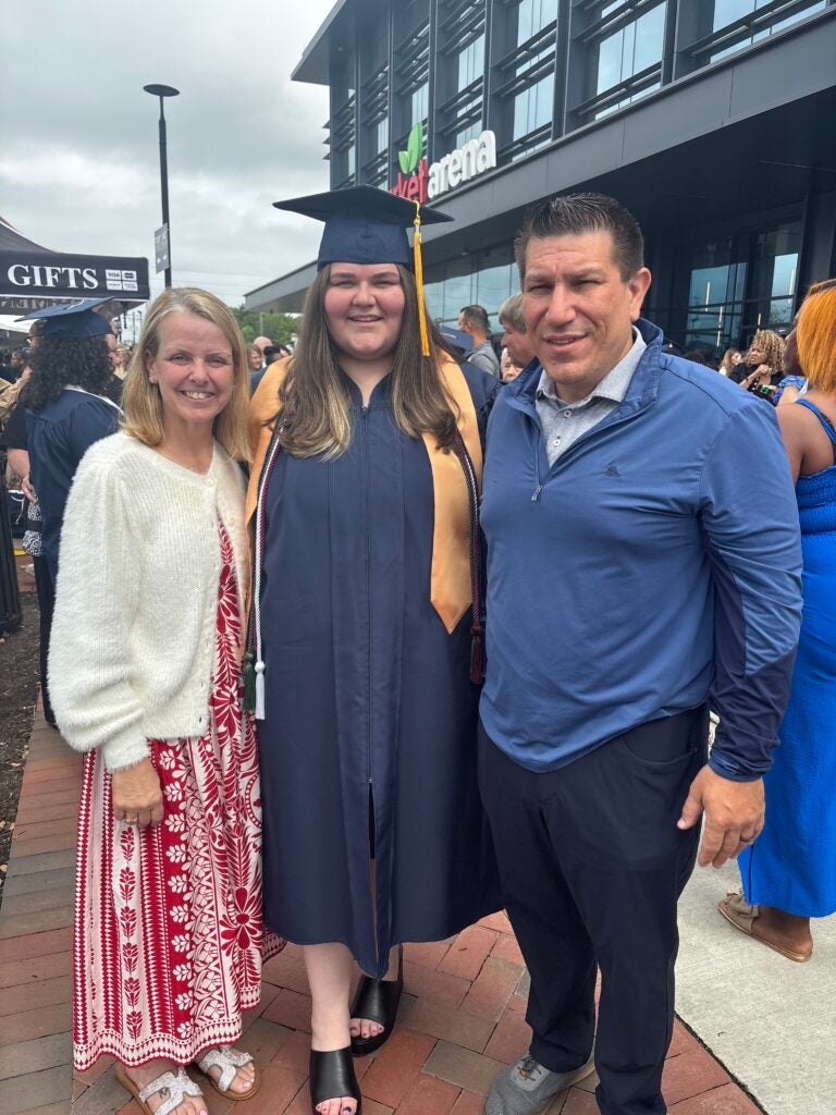 Graduate in navy cap and gown posing with two relatives outside a modern venue during graduation, all smiling.