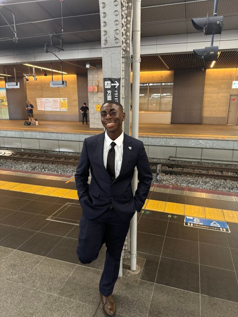 Smiling man in a nice black suit and black tie posing on a train platform with tracks behind him and a gray pole nearby.