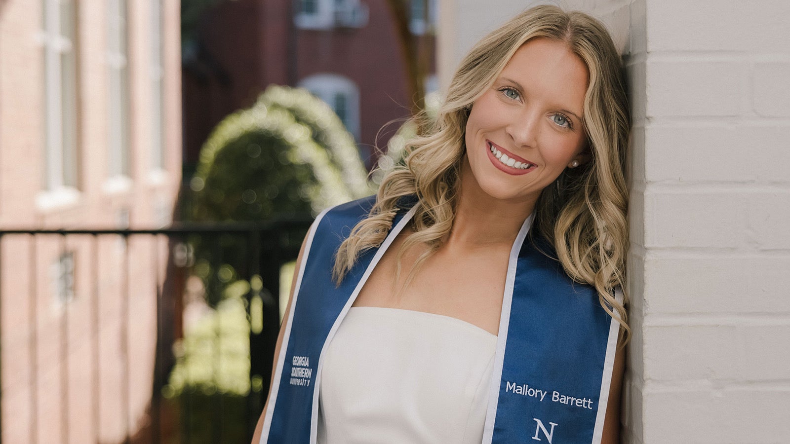 Smiling graduate Mallory Barrett in a white dress and blue sash leaning against a white brick wall.