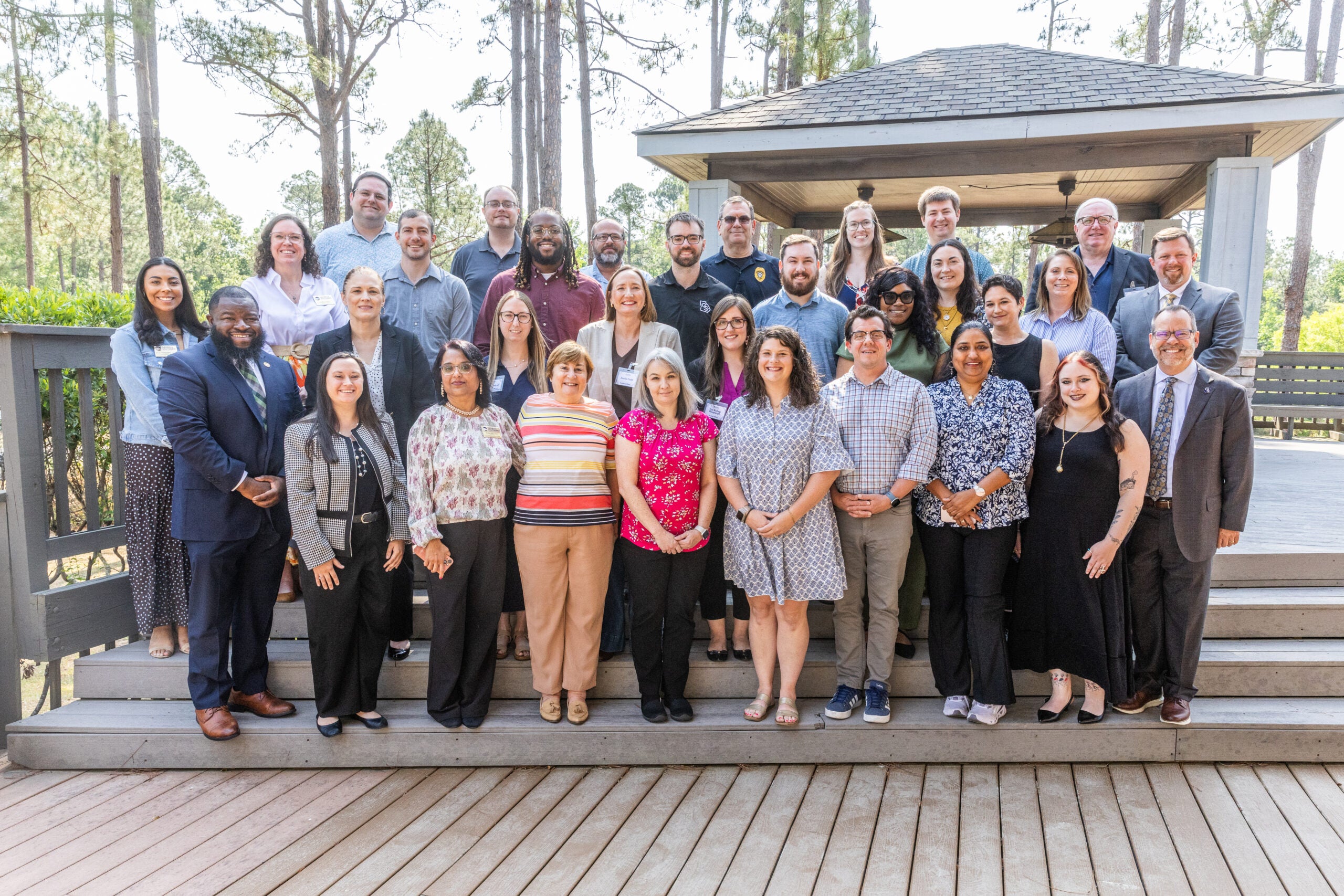 Large group of diverse adults standing on wooden steps outdoors for a group photo in a park setting. (About 25 people, smiling.)