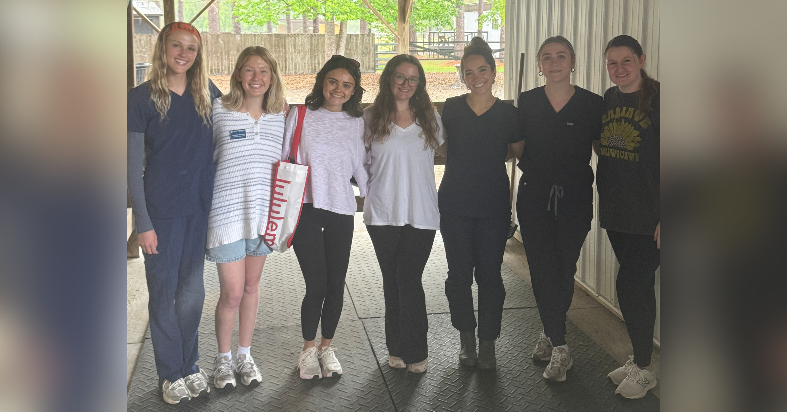 Group of seven Georgia Southern Communications Sciences and Disorders students standing side by side under a covered outdoor structure, smiling at the camera. They have their arms around each other in a friendly pose. The setting appears to be a wooded or park-like area with trees, a wooden fence, and picnic tables visible in the background. The students are dressed casually, with a mix of scrubs, T-shirts, and sweaters, suggesting a relaxed, collegial atmosphere.