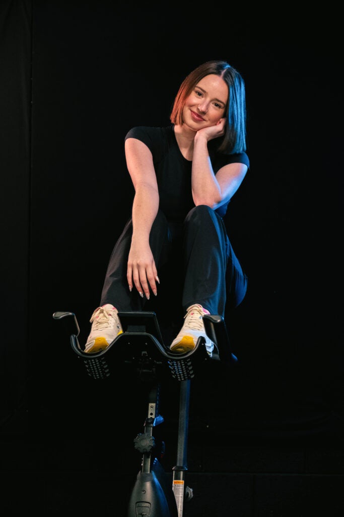 Woman sits on a stationary exercise bike in a dark studio, resting her chin on her hand and smiling slightly, with blue lighting on her hair and shoulders.