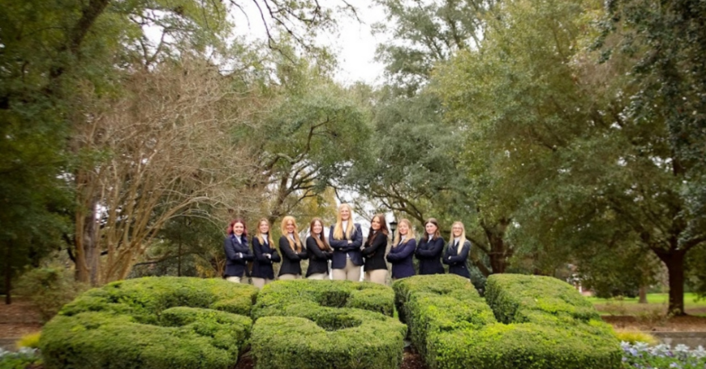 Group of young women in navy blazers posing with arms crossed in a park with trimmed hedges.
