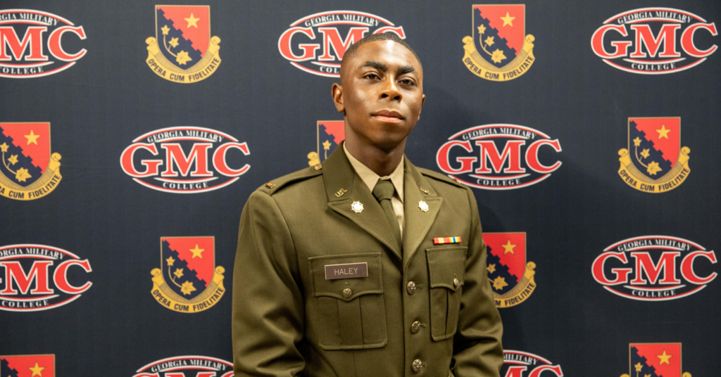 Man in a green dress uniform posing in front of a GMC College backdrop with logos and crests behind him, looking at the camera.