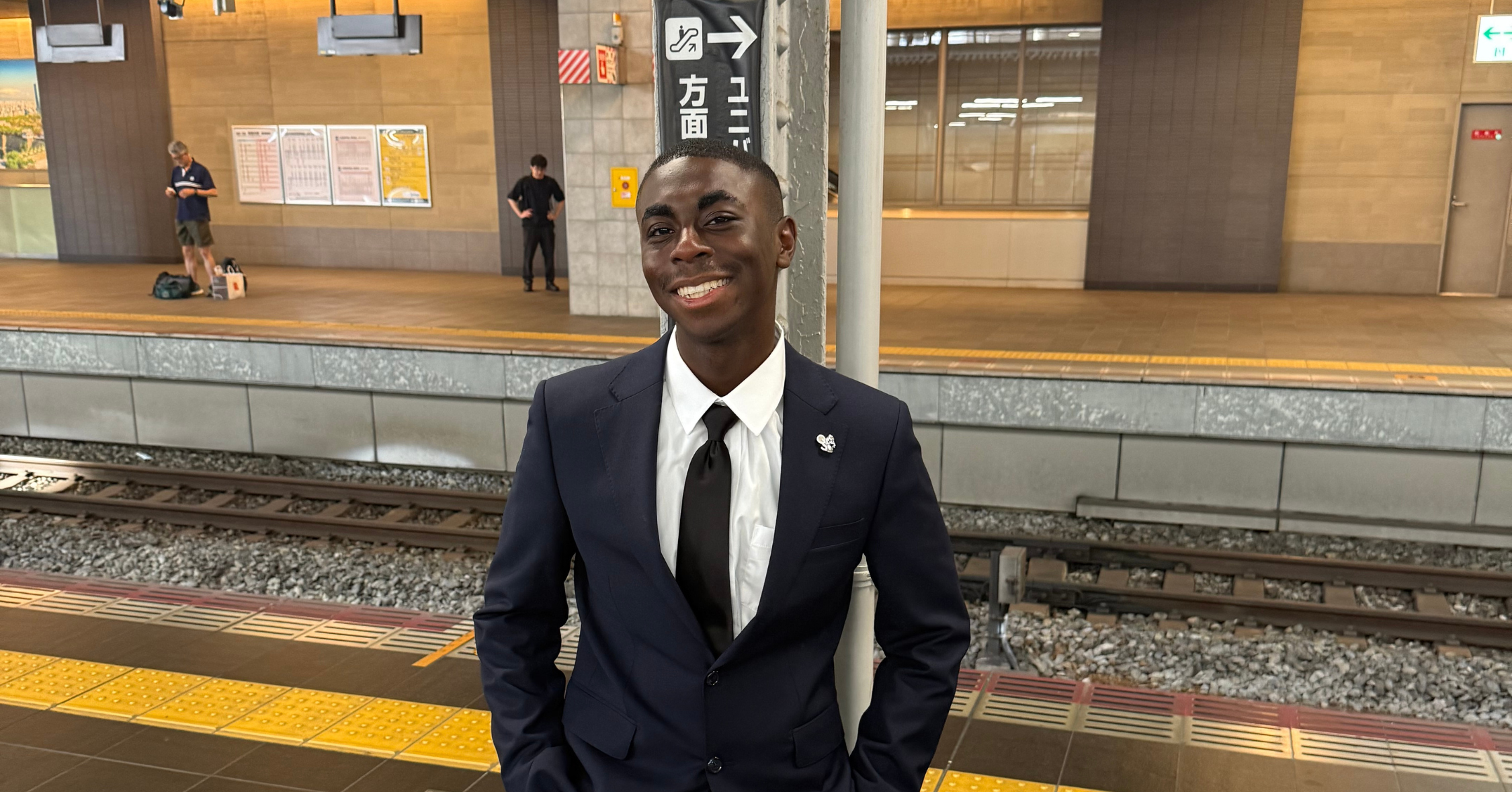 A person in a nice dark suit and tie with short black hair stands smiling on a train platform with tracks and other people in the background.