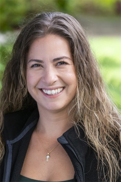Woman with long brown hair smiling outdoors, wearing a black jacket and gold cross necklace.