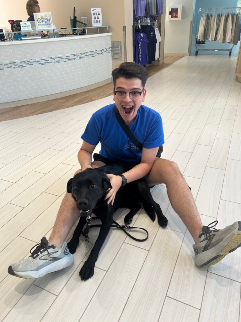 Xander Russell sits on the floor with a dog. He has an excited expression on his face.