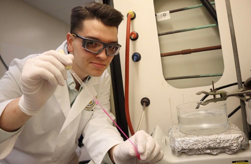 Xander Russell conducting research in a lab. He is wearing a variety of protective gear and using a pipette to handle a pink liquid.