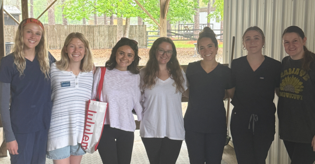 Group of seven Georgia Southern Communications Sciences and Disorders students standing side by side under a covered outdoor structure, smiling at the camera. They have their arms around each other in a friendly pose. The setting appears to be a wooded or park-like area with trees, a wooden fence, and picnic tables visible in the background. The students are dressed casually, with a mix of scrubs, T-shirts, and sweaters, suggesting a relaxed, collegial atmosphere.
