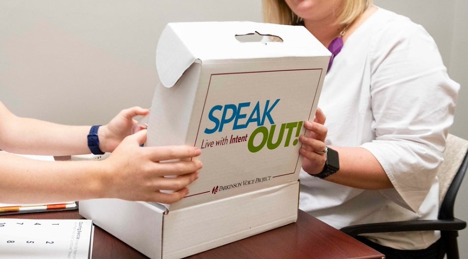 Person opening up white cardboard box with the words "SPEAK OUT" written across the outside of the box. Another person in a white shirt is also holding the box open.
