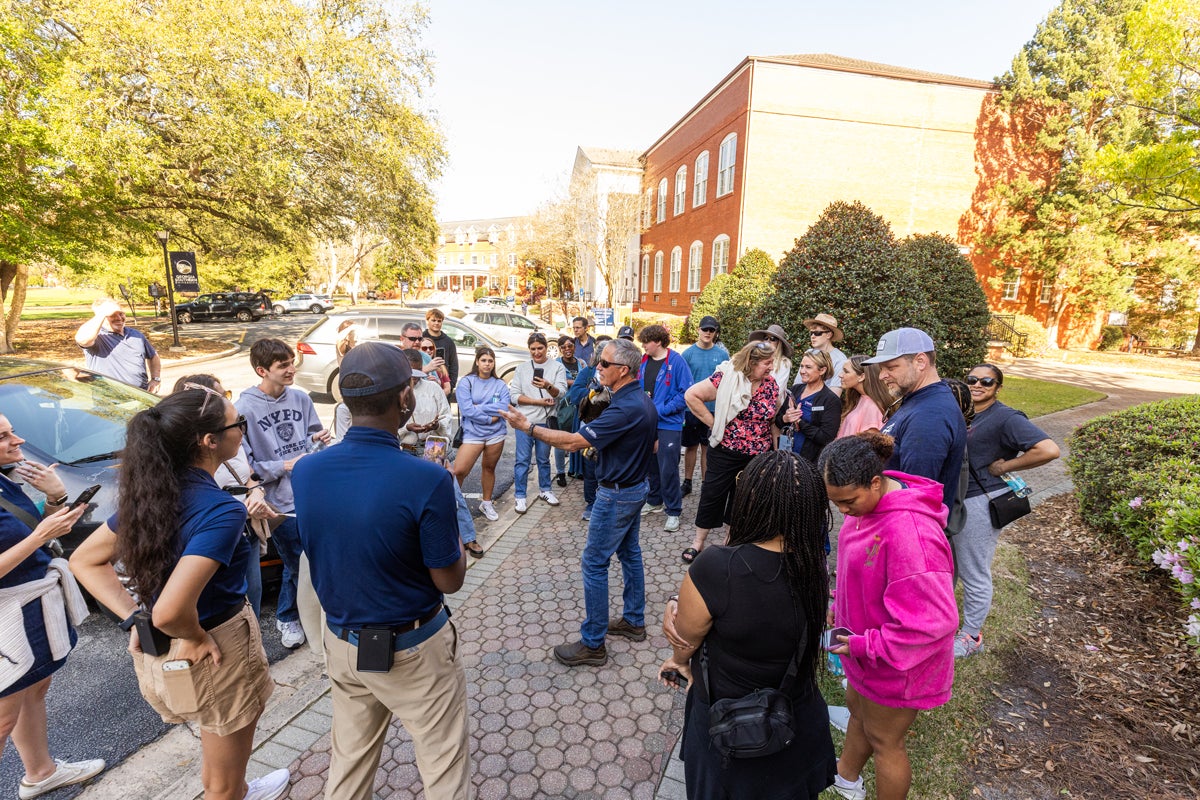 Group of diverse people listening to a man speaking on a sunny college campus path outside brick buildings.