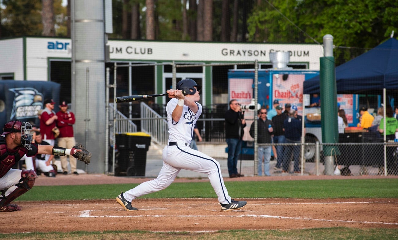 Baseball batter in a white uniform swings at a pitch at home plate, catcher crouched behind him.