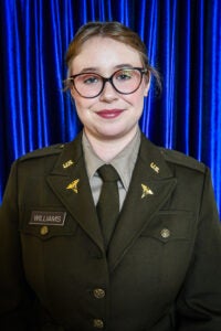 Female service member in olive U.S. Army dress uniform with name badge Williams, wearing glasses, standing in front of a blue curtain.