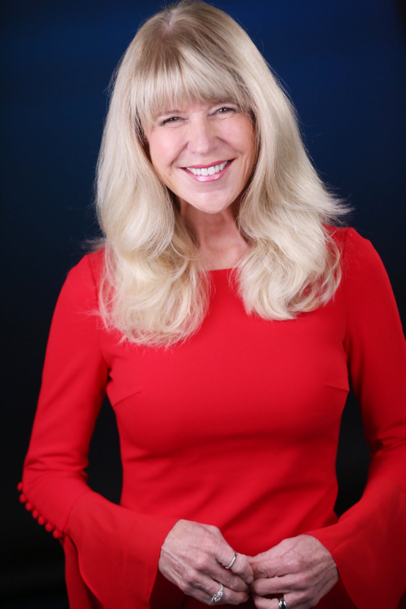 Smiling woman with long blonde hair in a red dress, hands clasped in front, dark background behind her.