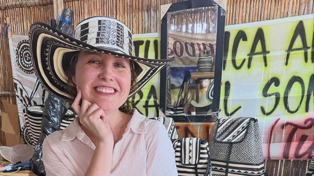 Woman smiling in a large black-and-white sombrero, seated among woven textiles and bright signs behind her.