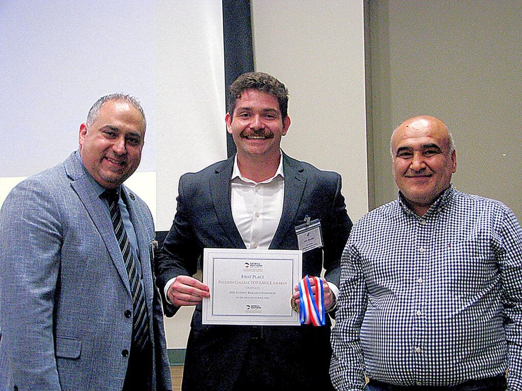 Thre people stand together and the middle person is holding an award certificate. They are standing on a stage in front of a screen.