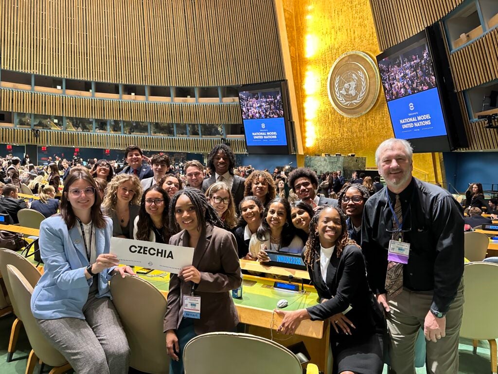 Diverse group of students taking a selfie in a UN conference hall, holding a Czechia sign.