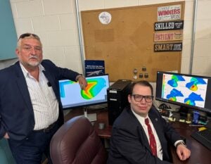 Brad Willis is sitting in a chair and Valentin Soloiu, Ph.D. is standing behind him. They are in a lab with two computer screens showing energy models.