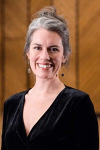 Smiling woman with gray hair in a black V-neck top, standing against a wooden background with earrings visible.